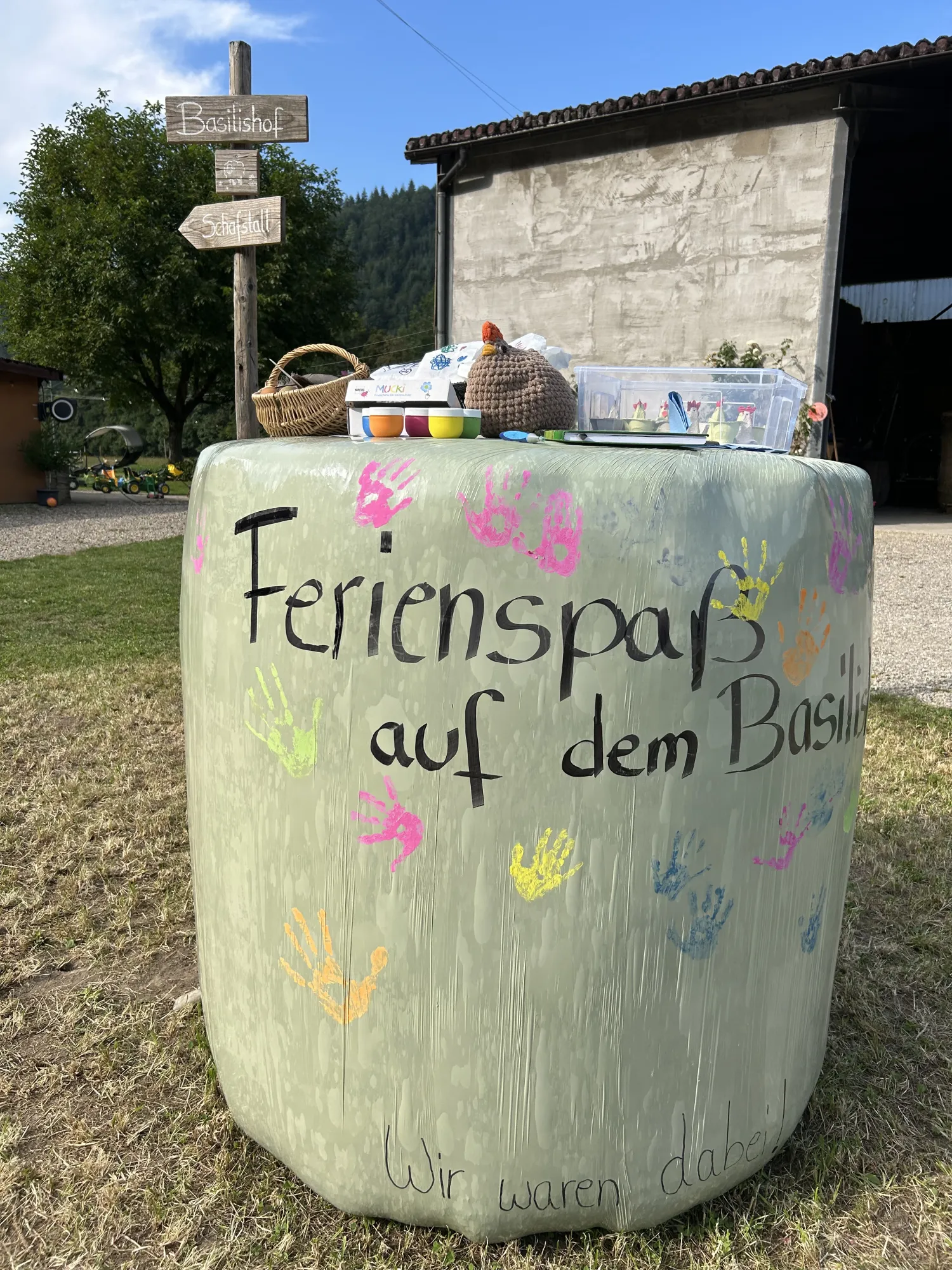 Eine bemalte Siloballen steht auf einer Wiese, dekoriert mit Handabdrücken und dem Schriftzug „Ferienspaß auf dem Basilishof“ - ein Angebot im Rahmen "Bauernhof erleben"