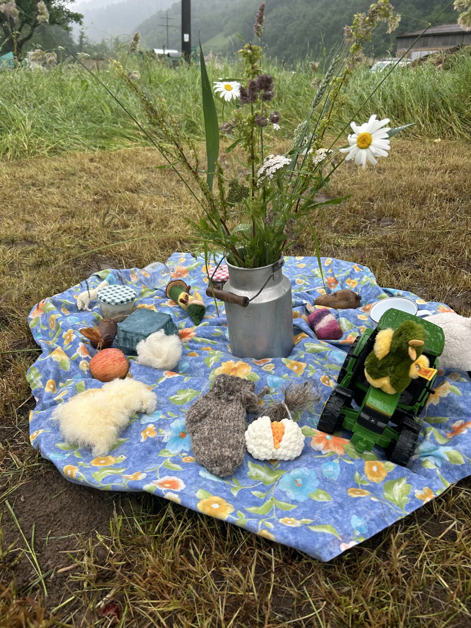 Ein Milchkrug mit Wiesenblumen steht auf einem Tuch, umgeben von kleinen Filztieren und einem Spielzeugtraktor.