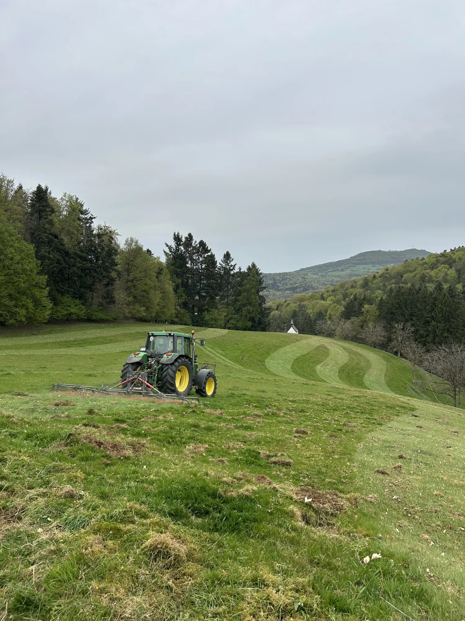 Ein Traktor arbeitet auf einer Hangwiese mit geschwungenen Mähstreifen, umgeben von Bäumen und Hügeln.