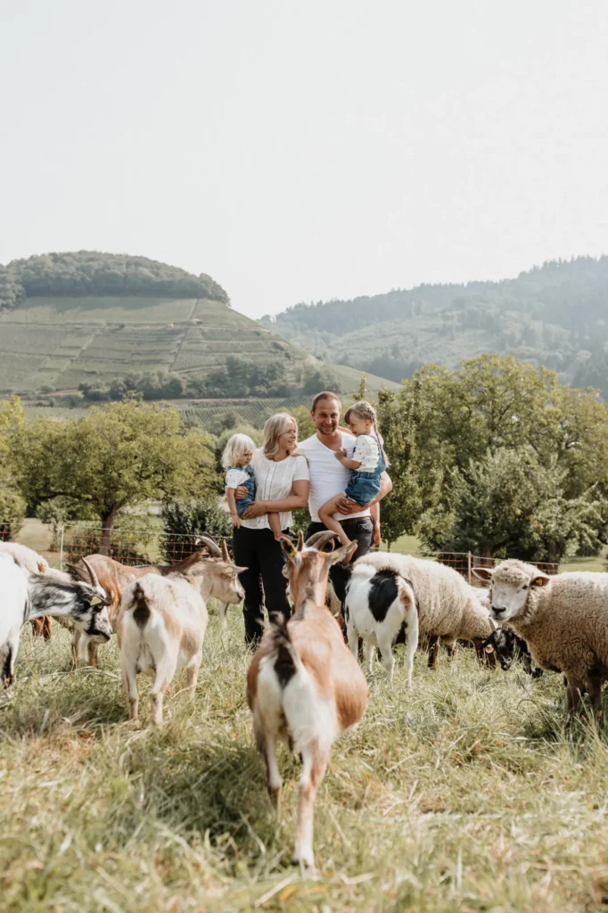 Zwei Erwachsene und zwei Kinder stehen auf einer Wiese zwischen Ziegen und Schafen.