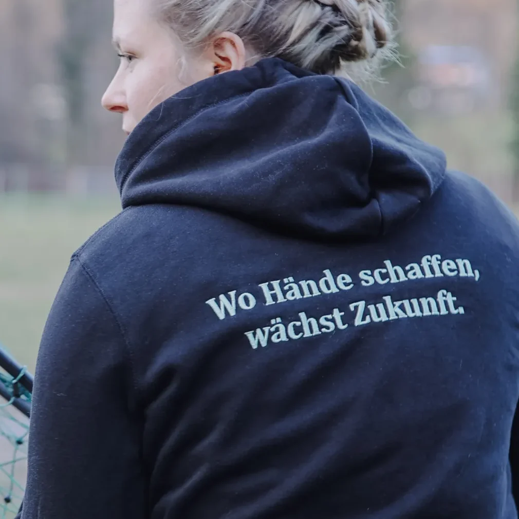 Eine Person hält ein grünes Netz, trägt einen dunklen Hoodie mit dem Rückenaufdruck „Wo Hände schaffen, wächst Zukunft“.